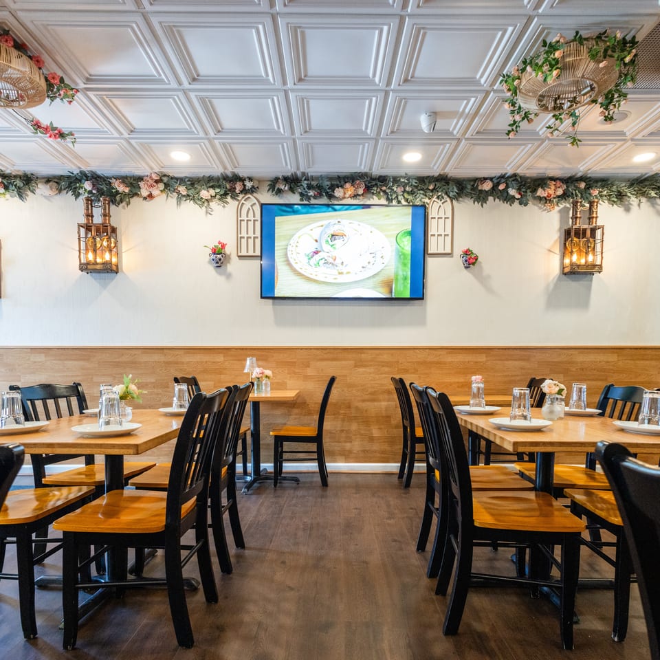 Restaurant dining room with wooden tables, black chairs, floral garlands, and a TV displaying food.