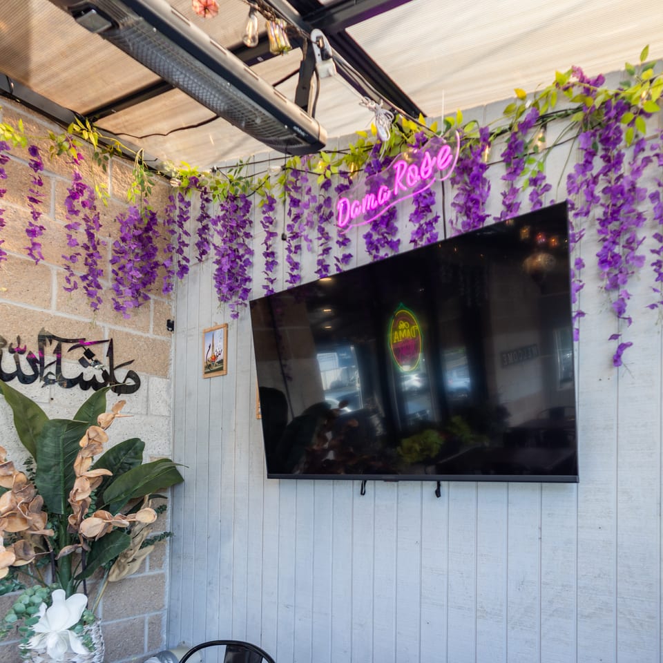 Purple wisteria decorates a restaurant wall with a "Dama Rose" neon sign, TV, and Arabic calligraphy.