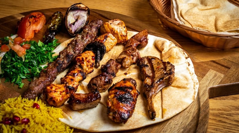 Grilled meat platter with kebabs, lamb chops, chicken, rice, salad, and flatbreads on a wooden board.