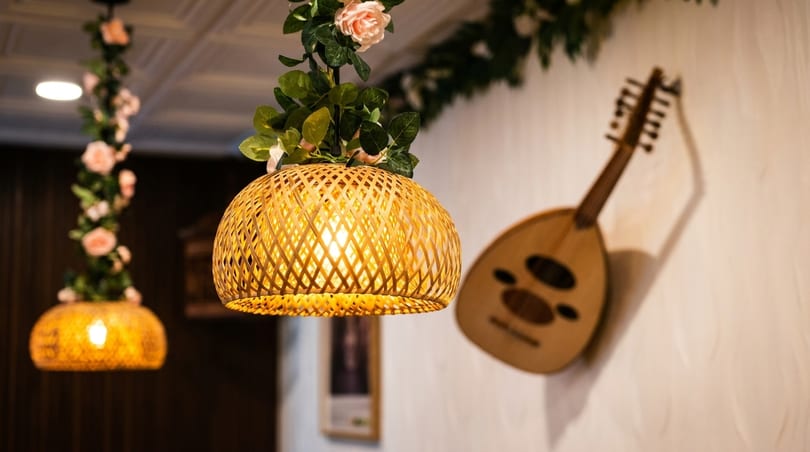 Warmly lit bamboo pendant lights adorned with pink flowers, beside a decorative wooden oud on a wall.