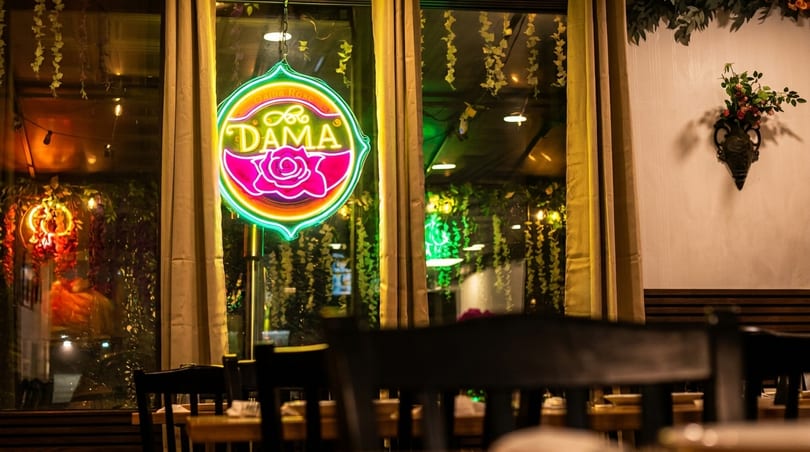 Warm restaurant interior with a vibrant "DAMA" neon sign, tables, and decorative plants.