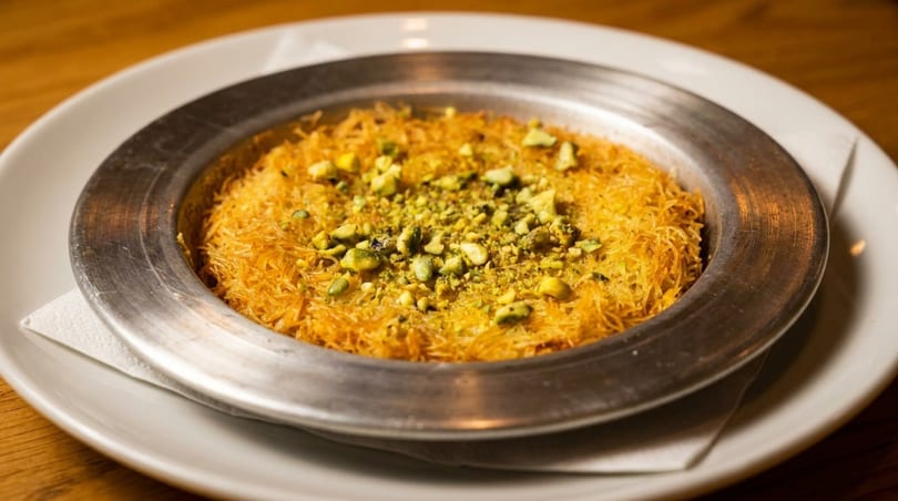 Golden kunefe dessert in a metal pan, topped with chopped pistachios, on a white plate.