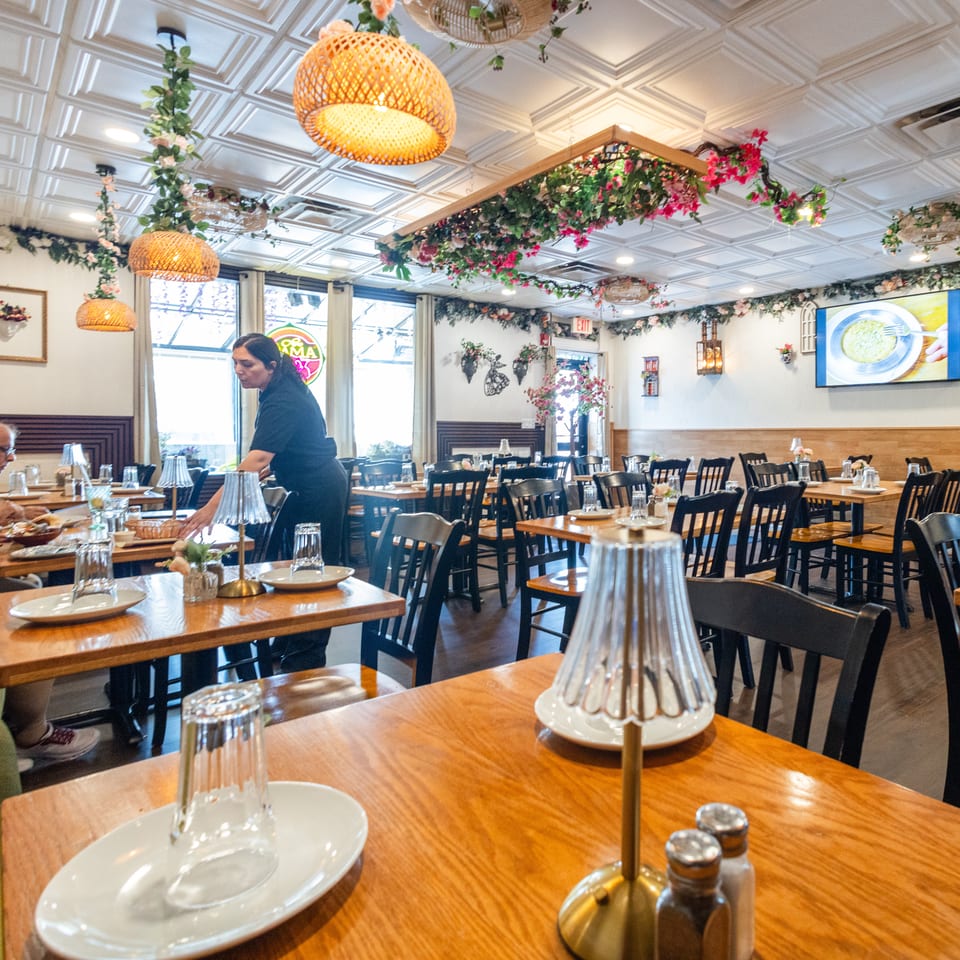 Restaurant interior with a waitress serving customers at wooden tables, featuring floral decor and warm lighting.