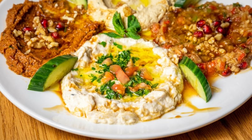 Platter of three Middle Eastern dips: hummus, muhammara, and baba ghanoush, garnished with fresh ingredients.