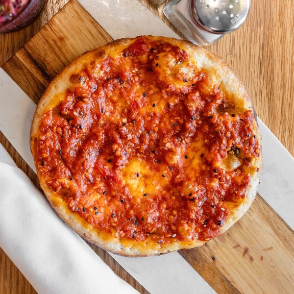 A round flatbread with vibrant red sauce and sesame seeds on a wooden serving board.