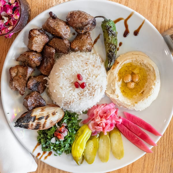 Grilled meat cubes, rice, hummus, grilled pepper, onion, and colorful pickled vegetables on a white plate.
