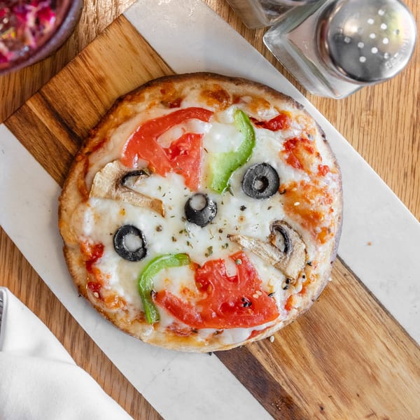 Personal pizza with tomatoes, green peppers, olives, and mushrooms on a marble and wood board.
