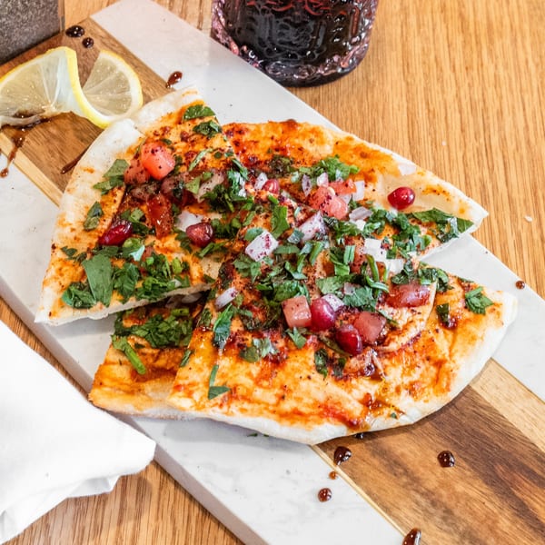 Flatbread slices with herbs, diced tomatoes, onions, and pomegranate seeds on a marble and wood board.