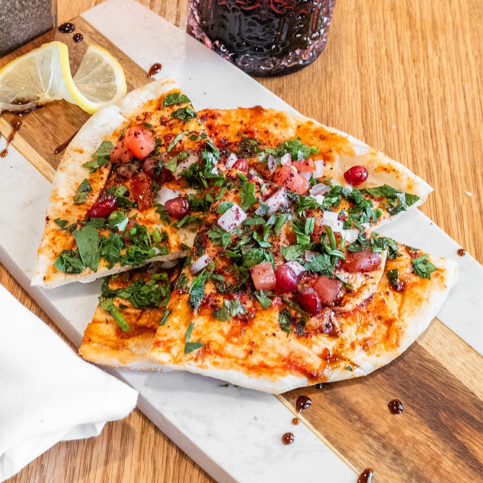 Flatbread slices with herbs, diced tomatoes, onions, and pomegranate seeds on a marble and wood board.