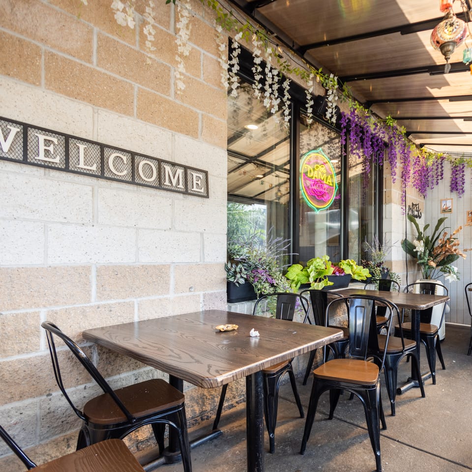 Outdoor restaurant patio with tables, chairs, a 'WELCOME' sign, and floral decorations.