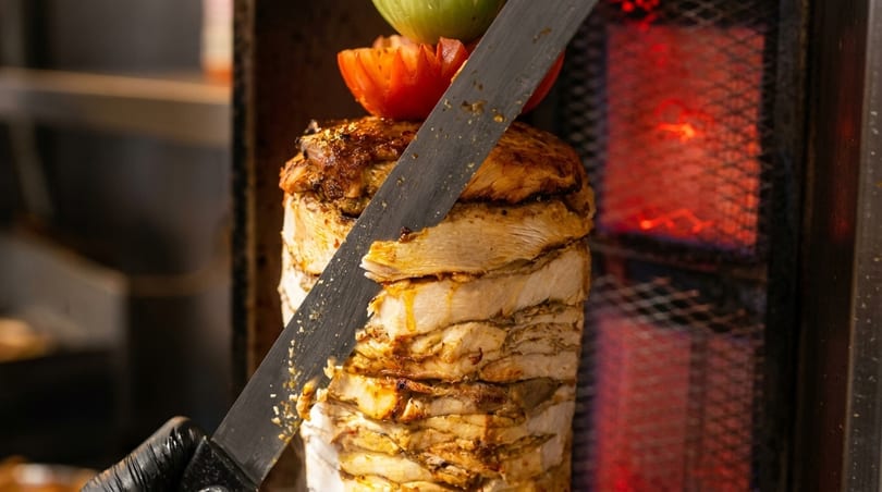 Slicing golden-brown chicken shawarma from a vertical spit, topped with tomato and green vegetable.