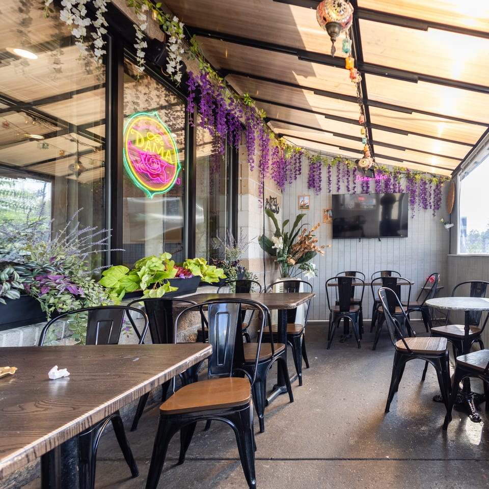 Inviting outdoor restaurant patio with black tables, purple flowers, and a bright "Dama" neon sign.