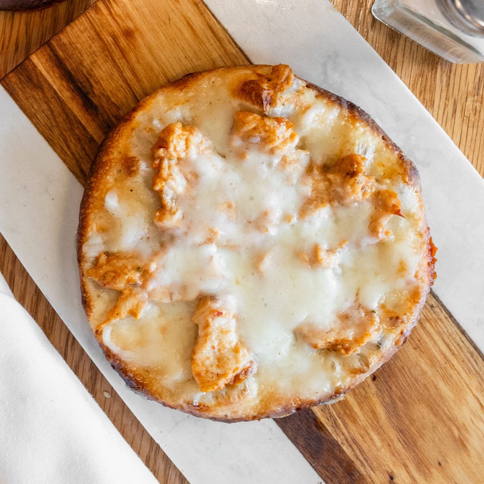 Overhead view of a small cheesy chicken flatbread on a striped wooden and marble serving board.