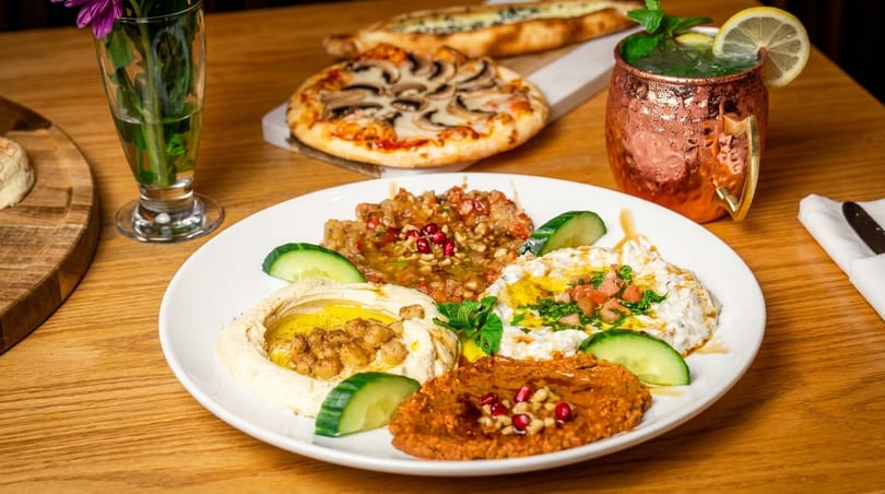 A mezze platter with hummus, baba ghanoush, and muhammara, alongside pizzas and a cocktail.