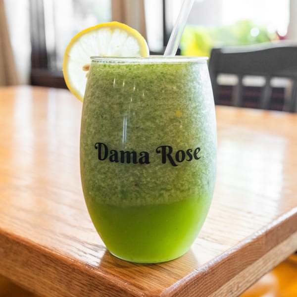 Green smoothie in a glass with "Dama Rose" text, lemon slice, and straw on a wooden table.