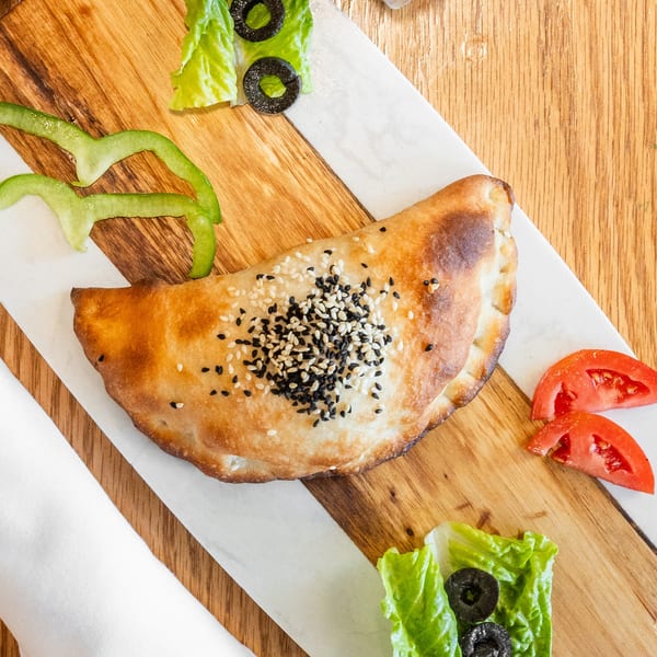Golden baked pastry with sesame seeds, garnished with bell pepper, lettuce, olives, and tomato on a serving board.