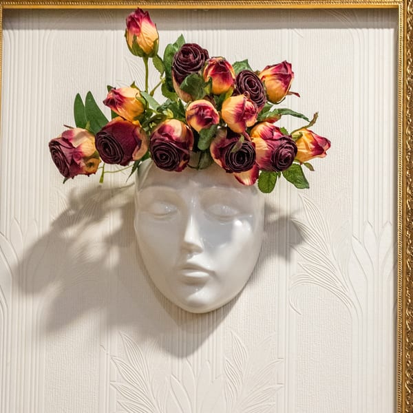 White ceramic face vase with burgundy and yellow roses, framed by a golden border on a textured wall.