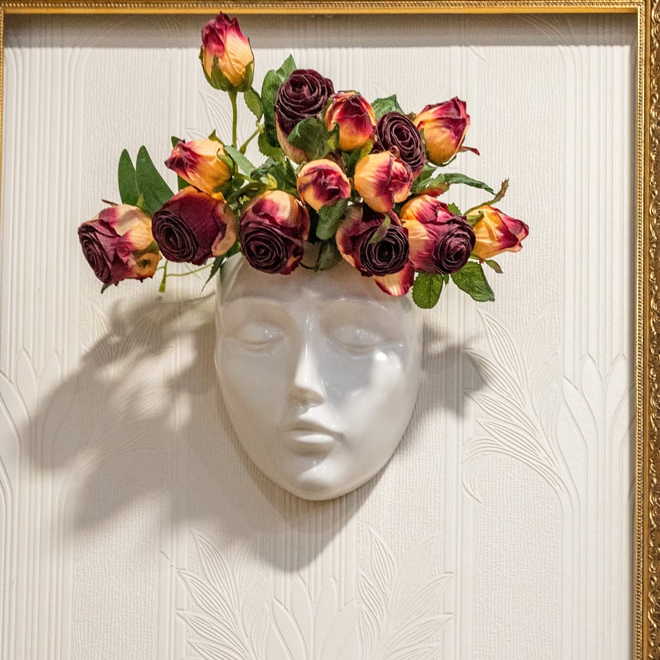 White ceramic face vase with burgundy and yellow roses, framed by a golden border on a textured wall.