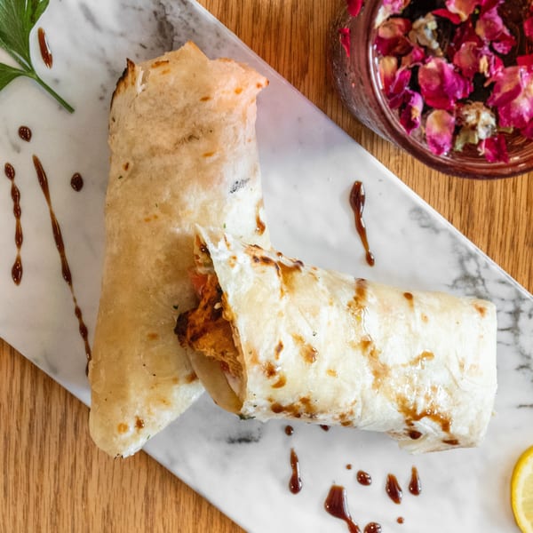 Two savory wraps with sauce, parsley, and lemon on a marble plate, with a rose petal drink.