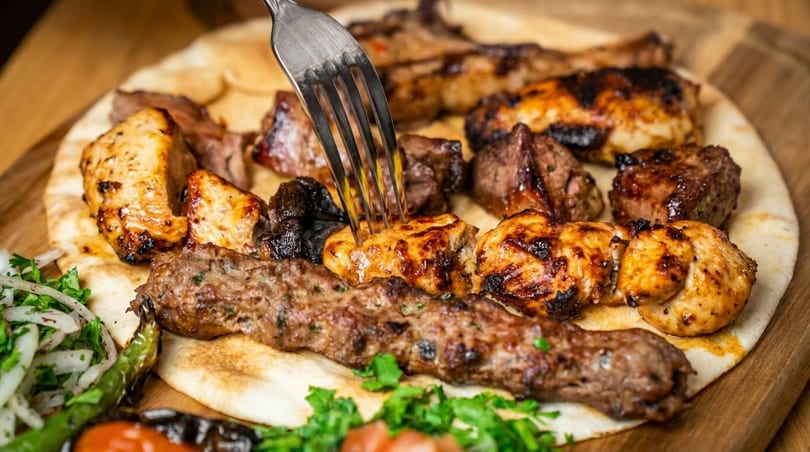 A fork piercing a piece of grilled chicken on a platter of various kebabs and flatbread.